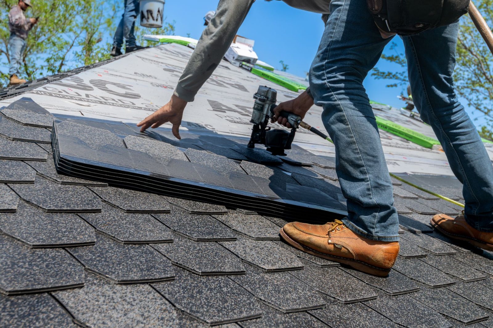 A harnessed roofer on a pitched multifamily roof laying a course of shingle with a nail gun, shot at distance from behind so the roof plane and the community below fill the frame.